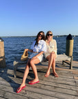 Two women sitting on a wooden dock bench with water and blue sky in the background. One wears a white shirt and shorts with red flip-flops and holds a tan sandal, while the other wears a cream lace-up top and shorts.