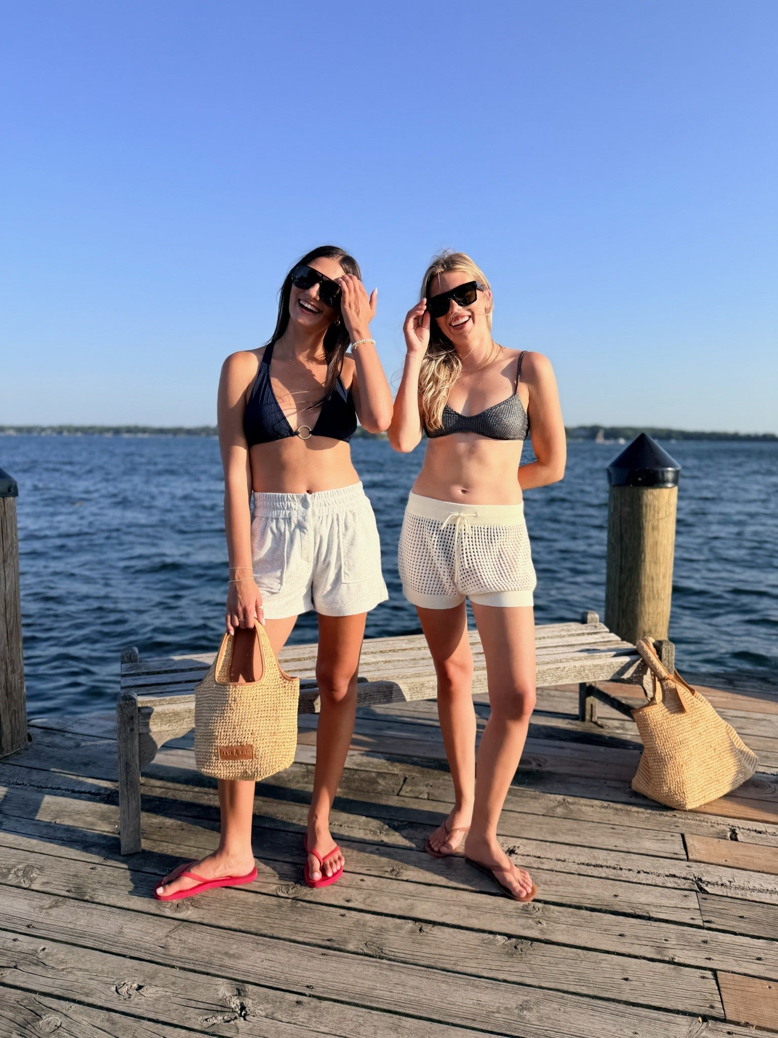 Two women posing on a wooden dock by the water, wearing matching bikini tops and white shorts. One wears a black bikini top and light gray shorts, while the other wears a gray bikini top and cream shorts.
