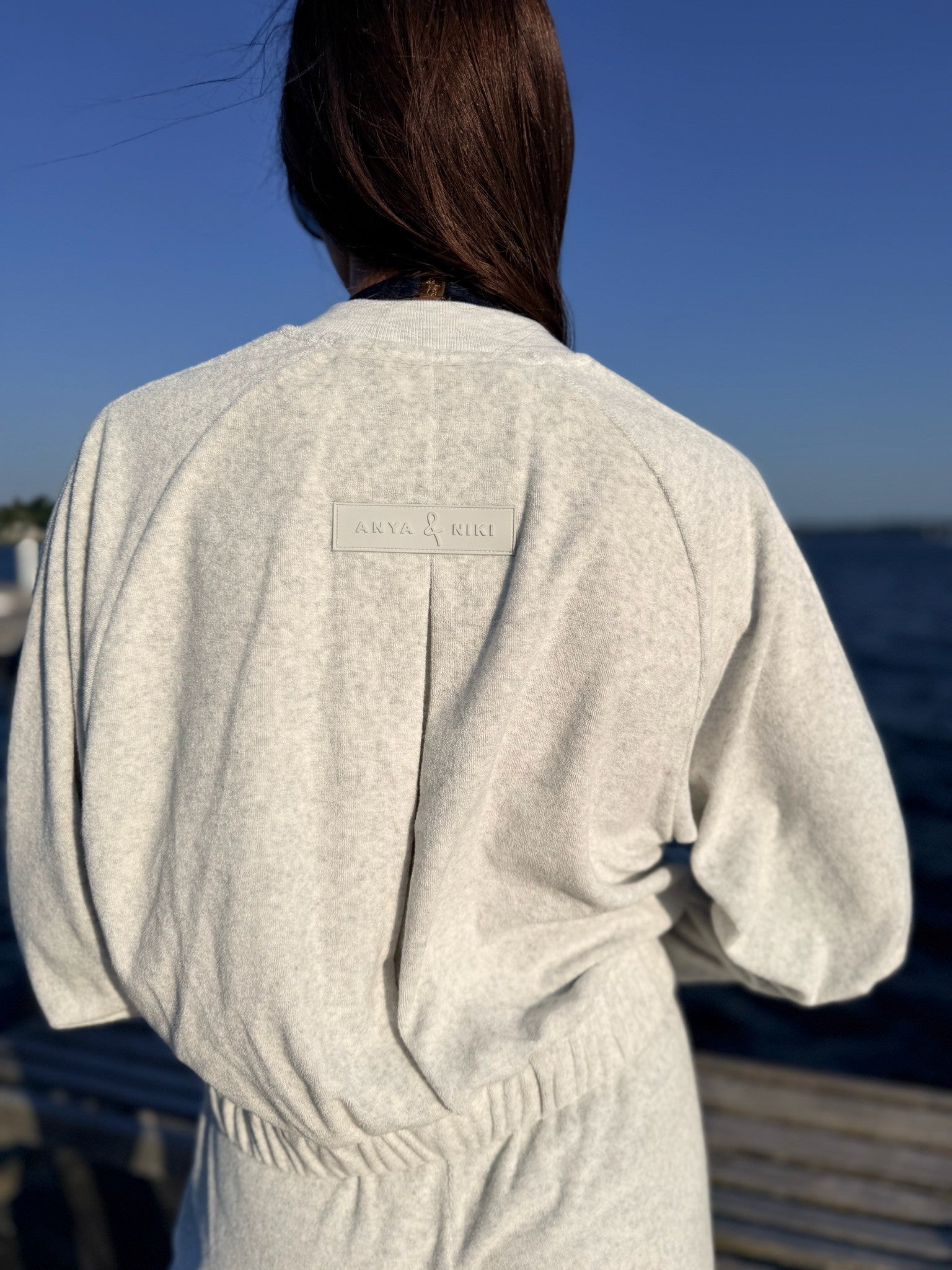 A person viewed from behind wearing a light heather gray sweatshirt with a small rectangular brand patch on the upper back, standing outdoors near water under a blue sky.