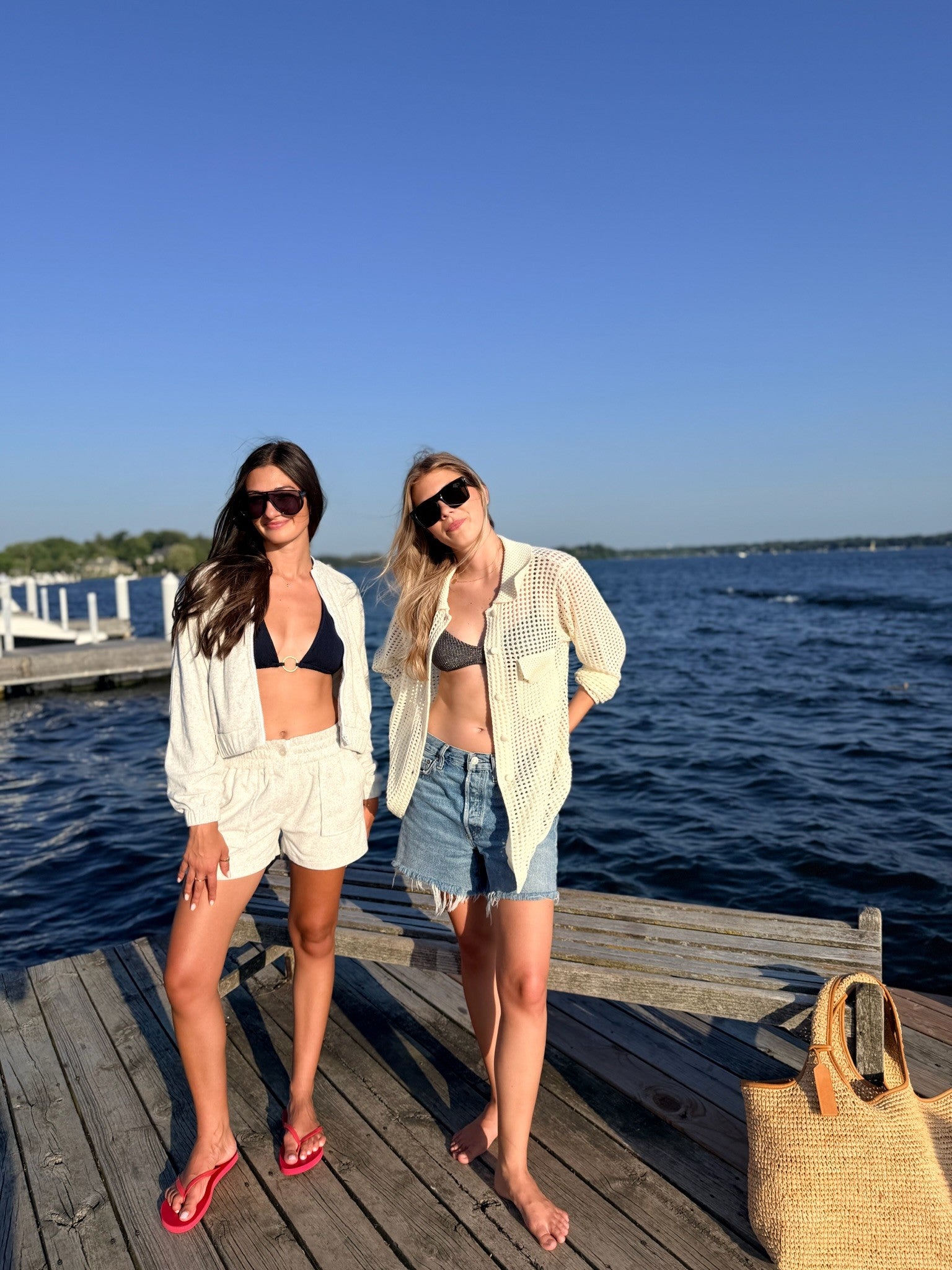 Two women posing on a wooden dock next to a body of water under a clear blue sky. One is wearing a white open-front jacket and grey shorts over a black bikini top, and the other is wearing a white open-front shirt and denim shorts over a gray bikini top.