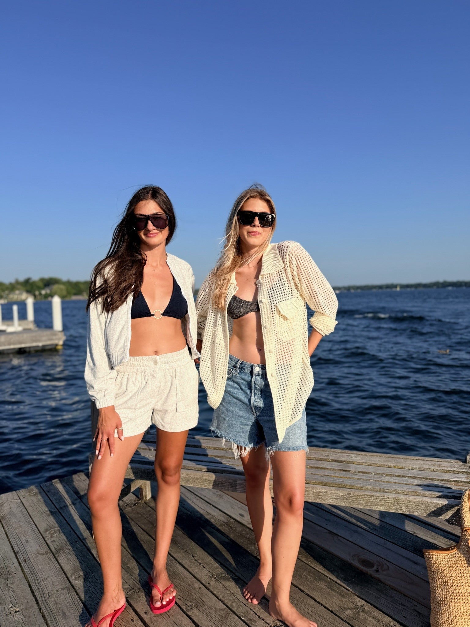 Two women standing on a wooden dock by the water wearing swimwear and open lightweight cover-ups under a clear blue sky.