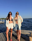Two women standing on a wooden dock by the water wearing swimwear and open lightweight cover-ups under a clear blue sky.