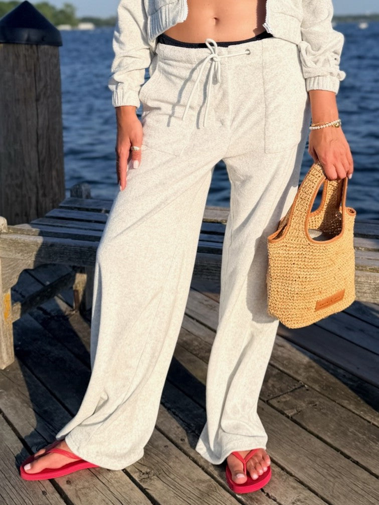 A person standing on a wooden dock by the water wearing light heather gray wide-leg drawstring sweatpants, a white cropped long-sleeve top, and red flip-flops, while holding a woven straw tote bag.