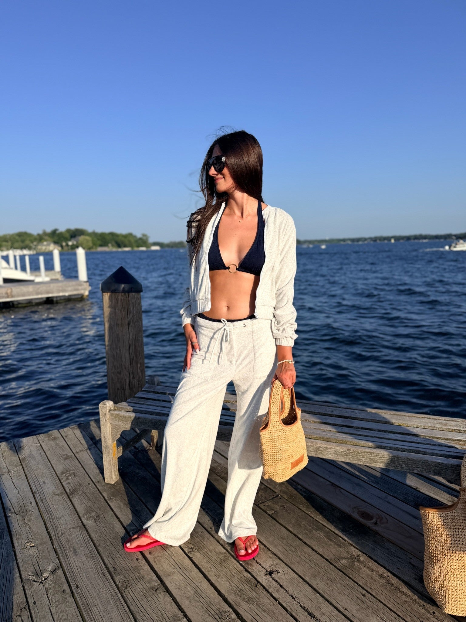 A woman stands on a wooden dock by the water wearing a light grey open-front jacket and matching wide-leg drawstring pants over a black bikini top, with red shoes and a straw tote bag.