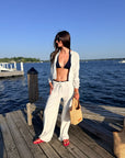 A woman stands on a wooden dock by the water wearing a light grey open-front jacket and matching wide-leg drawstring pants over a black bikini top, with red shoes and a straw tote bag.