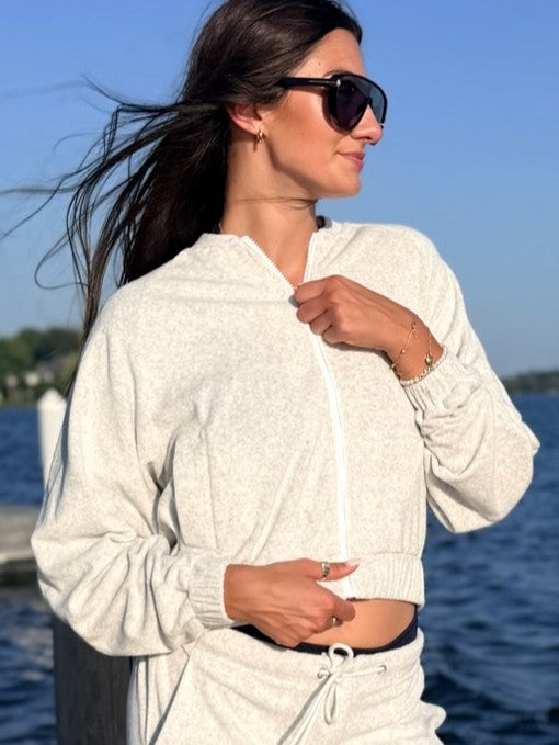 A woman wearing a light heather gray zip-up hoodie and matching drawstring pants stands outdoors by the water, wearing dark sunglasses and looking to the side.