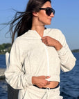A woman wearing a light heather gray zip-up hoodie and matching drawstring pants stands outdoors by the water, wearing dark sunglasses and looking to the side.
