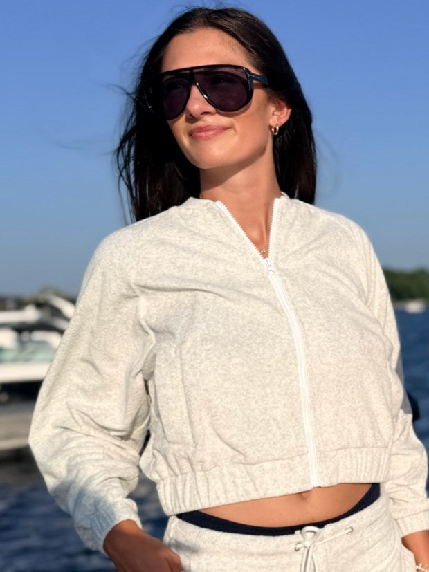 A woman wearing a light heather gray cropped zip-up jacket and matching sweatpants, styled with dark sunglasses, standing outdoors near water and boats.