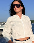 A woman wearing a light heather gray cropped zip-up jacket and matching sweatpants, styled with dark sunglasses, standing outdoors near water and boats.