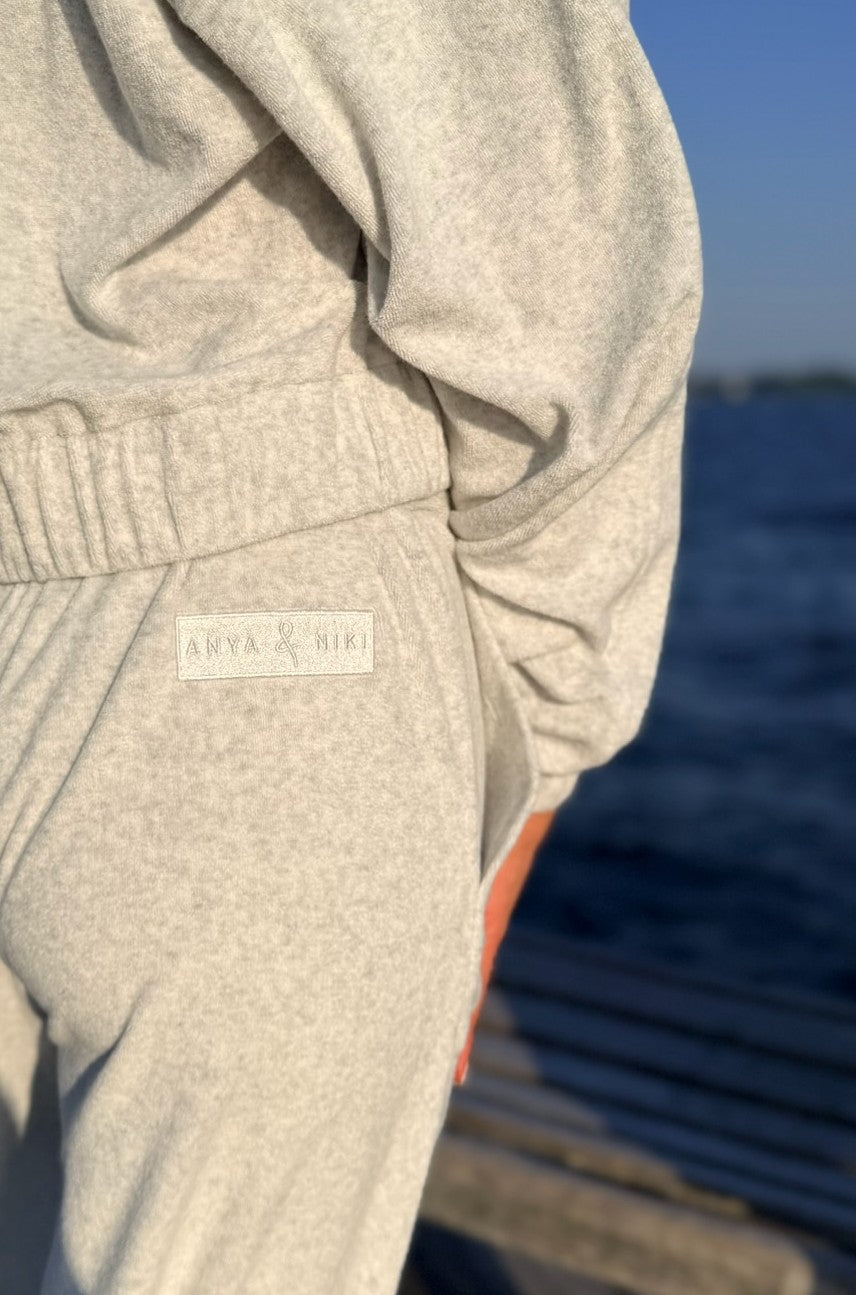 Close-up of a person wearing a light heather gray sweatshirt and matching jogger pants, with a small visible 'ANYA &amp; NIKI' label on the pocket area. The background shows an outdoor water scene with blue sky and blurred steps.