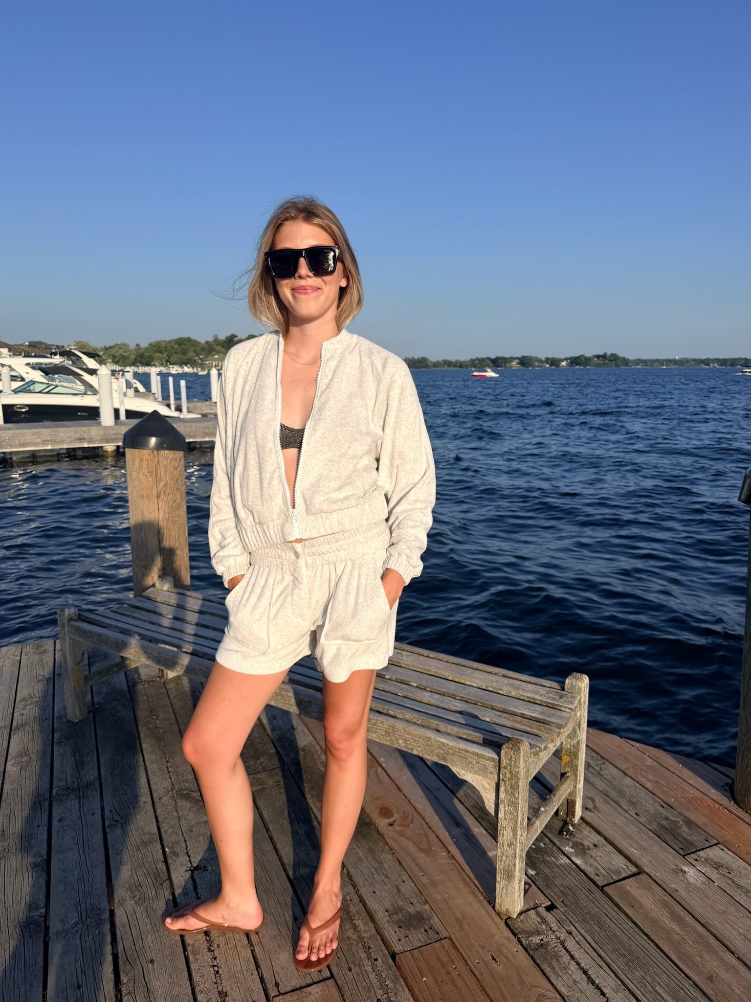 A woman wearing a light grey terry lounge set consisting of a long-sleeve button-up jacket and matching shorts, standing on a wooden dock with a lake and blue sky in the background.