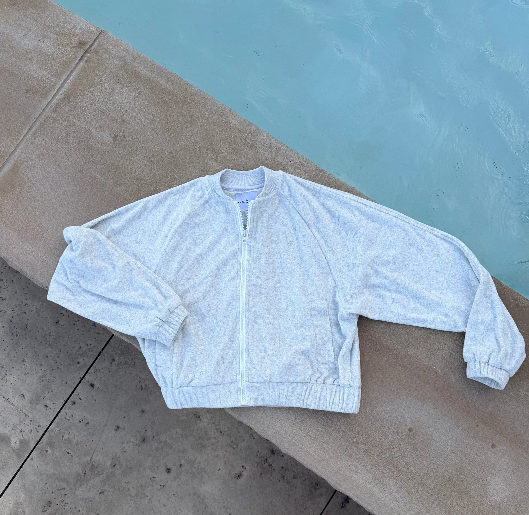 A light heather gray zip-up jacket with a stand collar and elastic cuffs, laid flat on a concrete poolside surface.