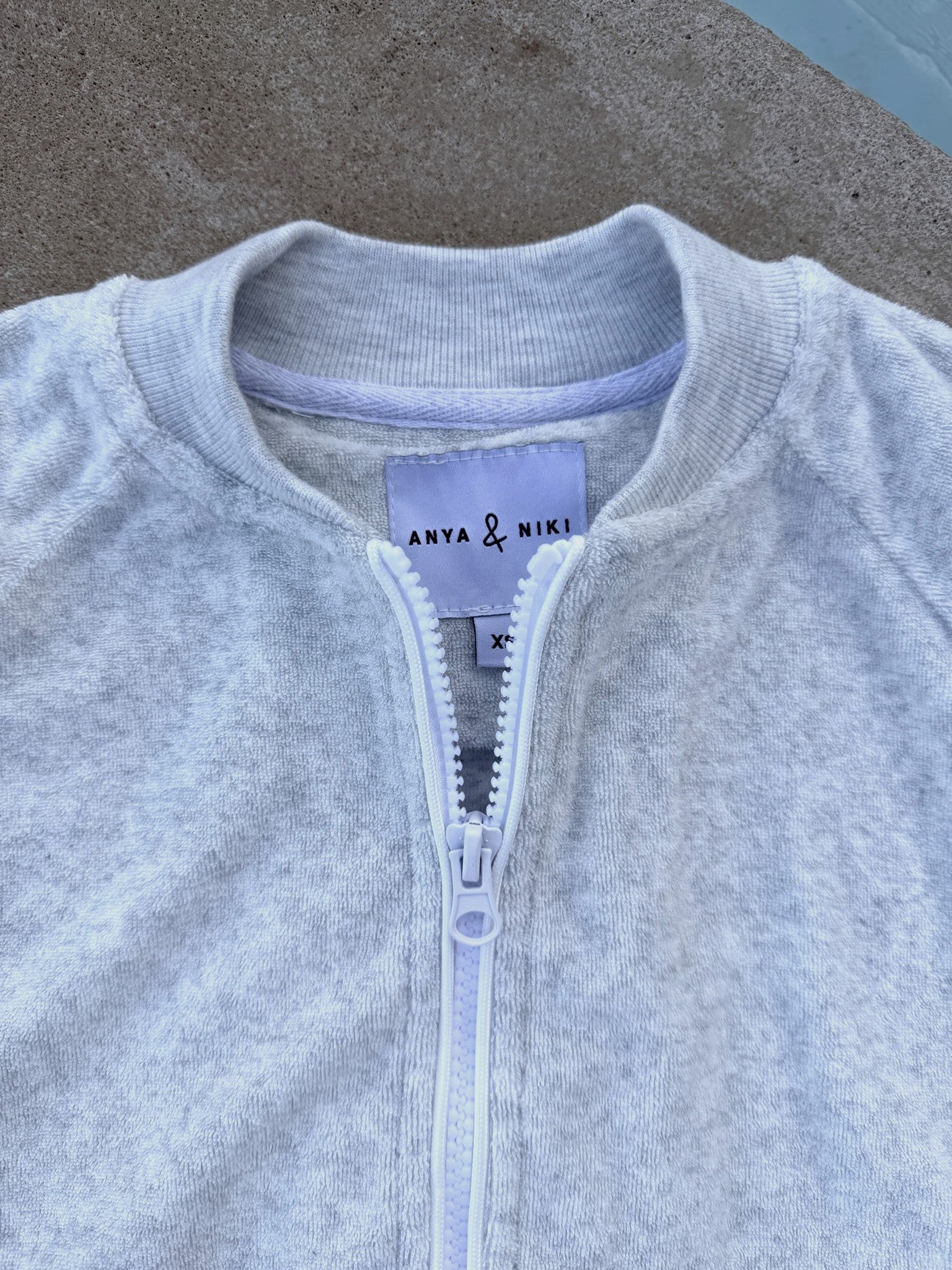 Close-up of a light heather gray zip-up sweatshirt with a ribbed collar, featuring a white zipper and a visible 'ANYA & NIKI' brand label.