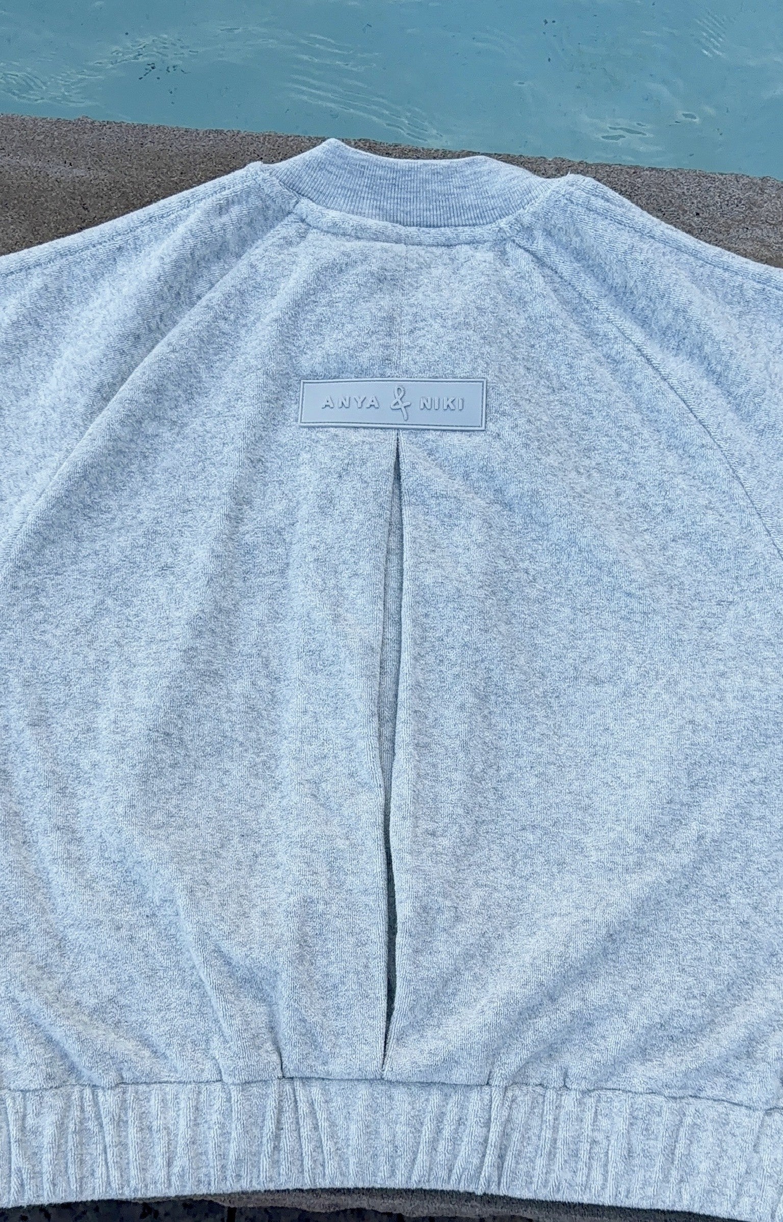 A light heather gray sweatshirt laid flat on a concrete surface with a swimming pool in the background. The back of the sweatshirt features a central seam and a small rectangular brand label near the collar.