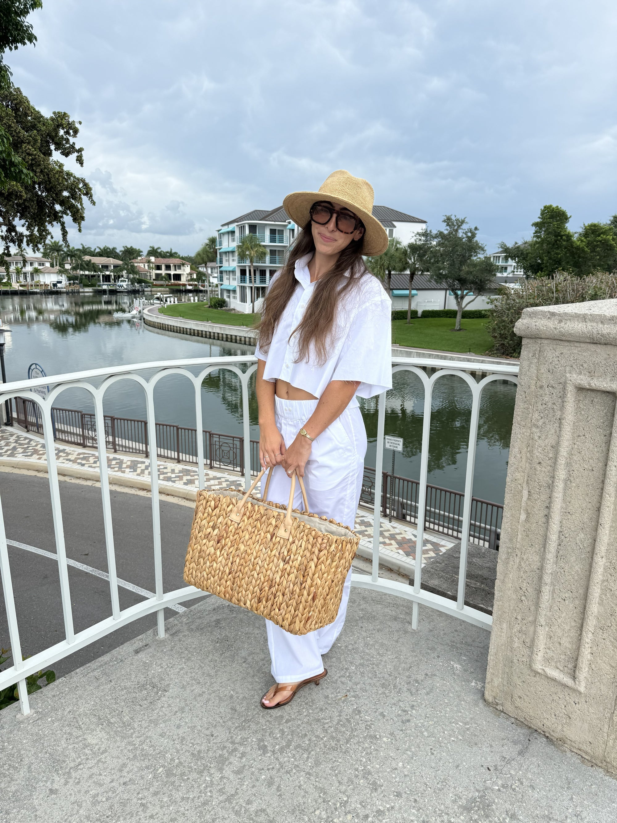 A woman stands on an outdoor balcony overlooking a canal, wearing a white short-sleeve crop top and white wide-leg pants, a beige straw hat, and holding a large woven straw tote bag.