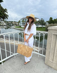 A woman stands on an outdoor balcony overlooking a canal, wearing a white short-sleeve crop top and white wide-leg pants, a beige straw hat, and holding a large woven straw tote bag.
