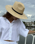 A woman wearing a wide-brimmed straw hat with a black band, a white short-sleeve button-up shirt, and white pants, standing outdoors near a railing with water and houses in the background.