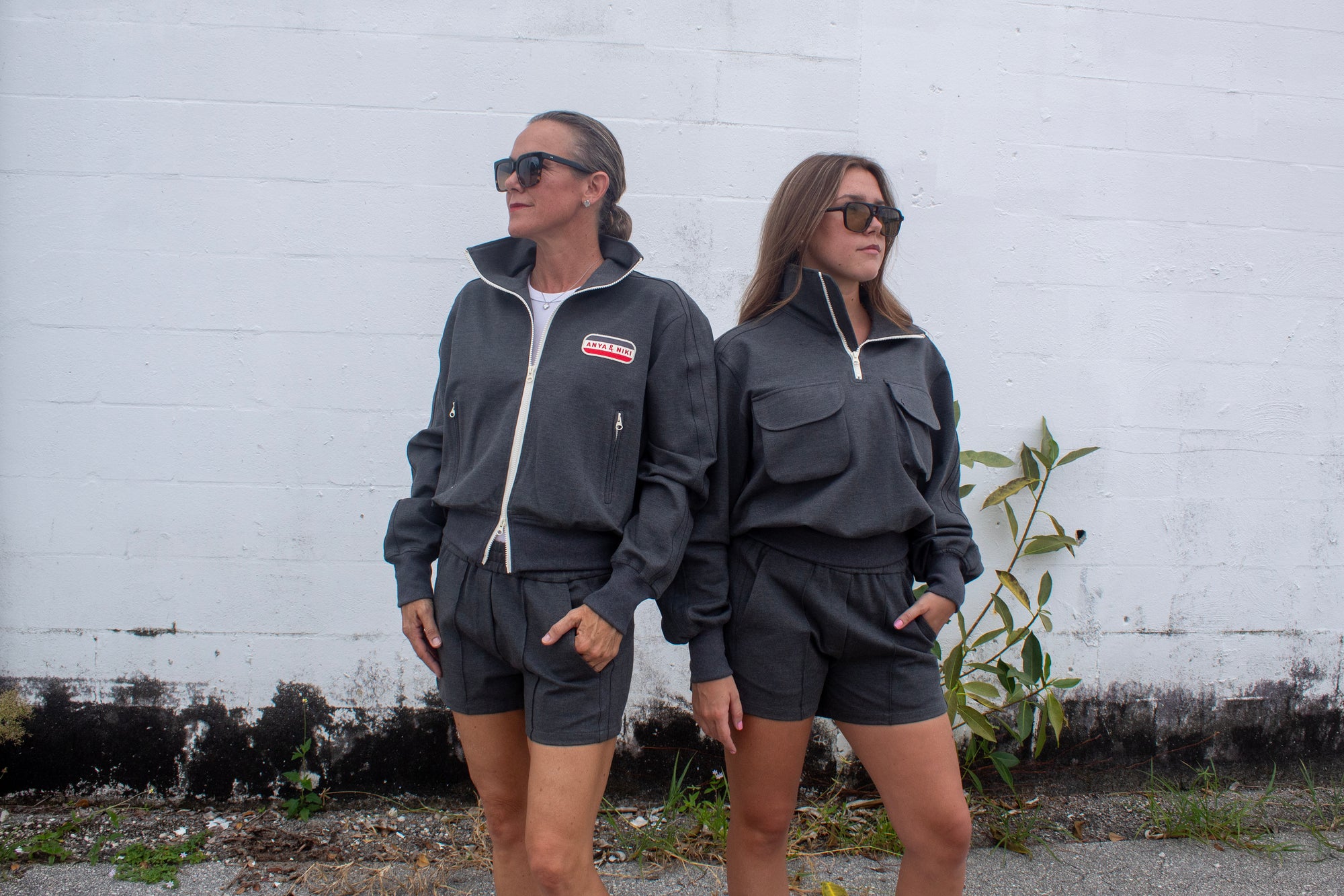 Two women wearing matching gray outfits standing against a white wall.
