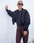 Person wearing a black jacket and brown pants against a white wall