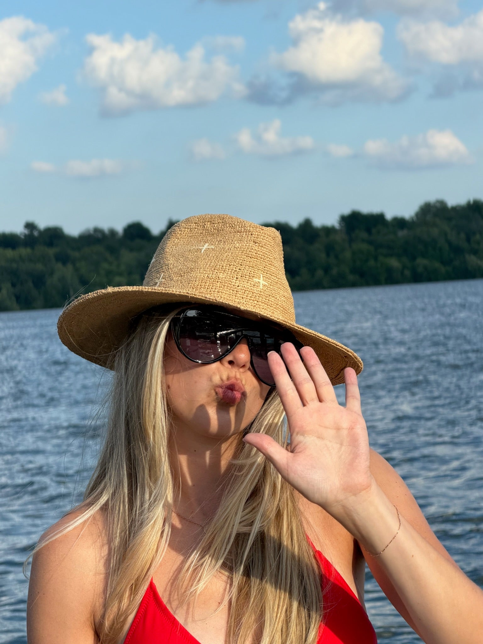 A woman with blonde hair wearing a straw sun hat, dark sunglasses, and a red top poses in front of a lake with trees and a blue sky in the background.