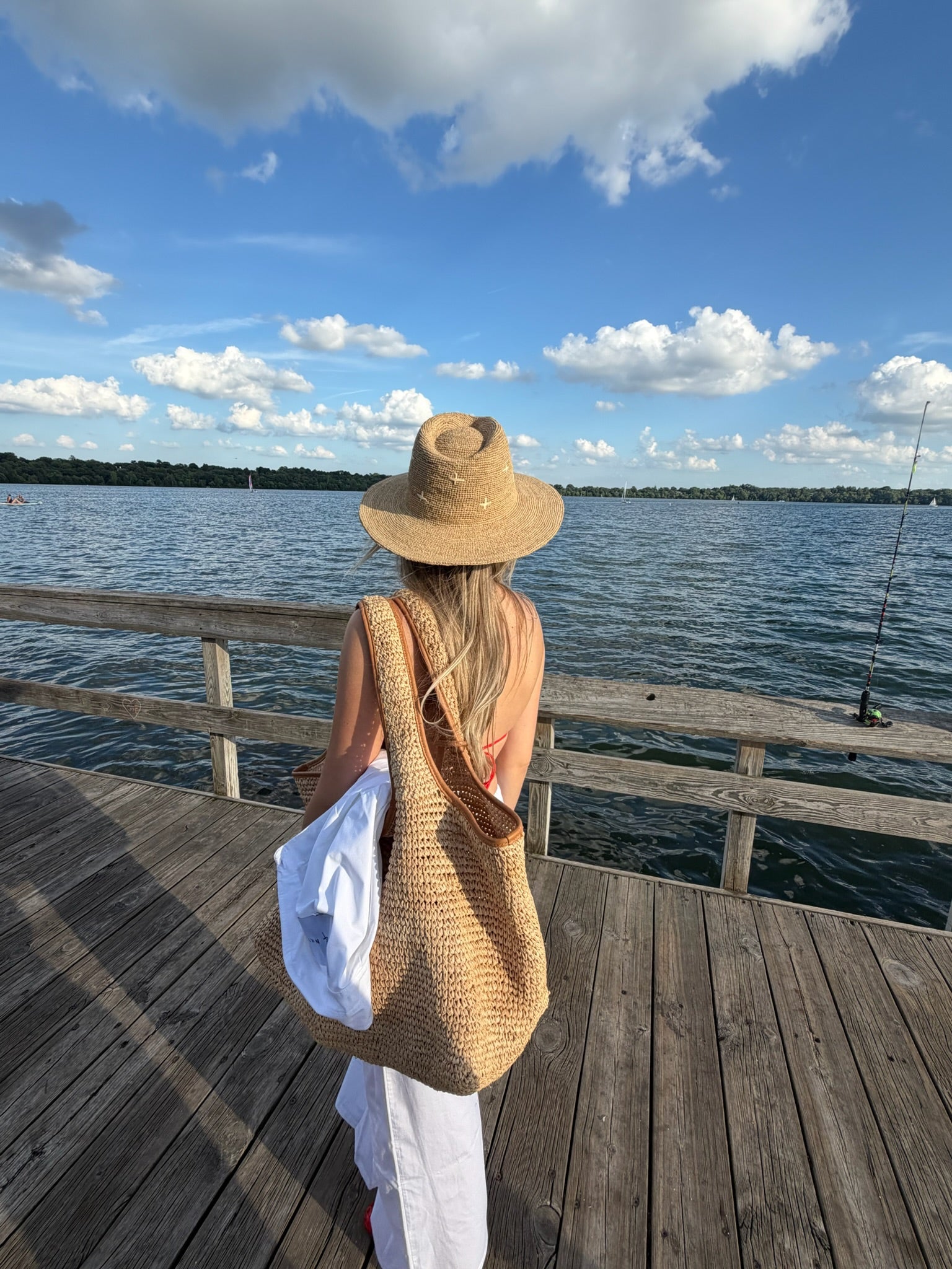 A woman viewed from behind on a wooden dock by the water, wearing a wide-brim straw hat, a large woven straw shoulder bag, and white pants, with a white shirt tucked into the bag.