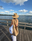 A woman viewed from behind on a wooden dock by the water, wearing a wide-brim straw hat, a large woven straw shoulder bag, and white pants, with a white shirt tucked into the bag.