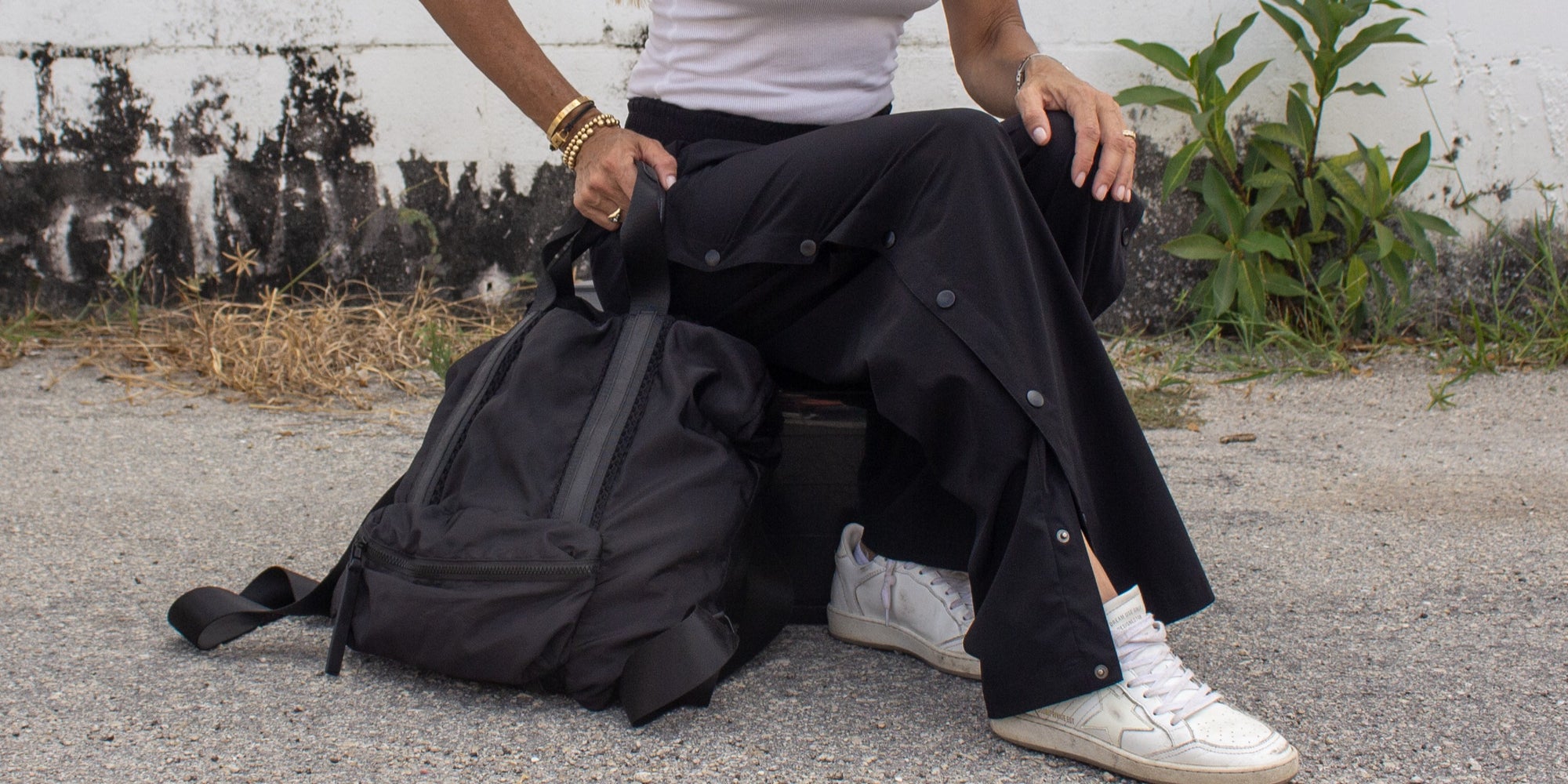 Person sitting by a black backpack wearing a white shirt and black pants with white sneakers.