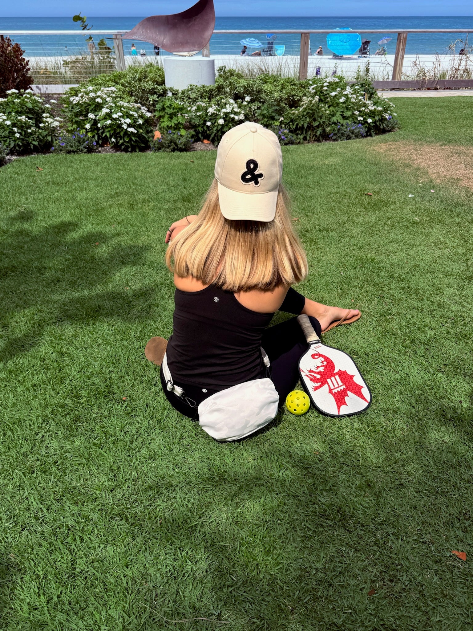 Person sitting on grass with a paddle and ball, wearing a cap with a black ampersand logo.