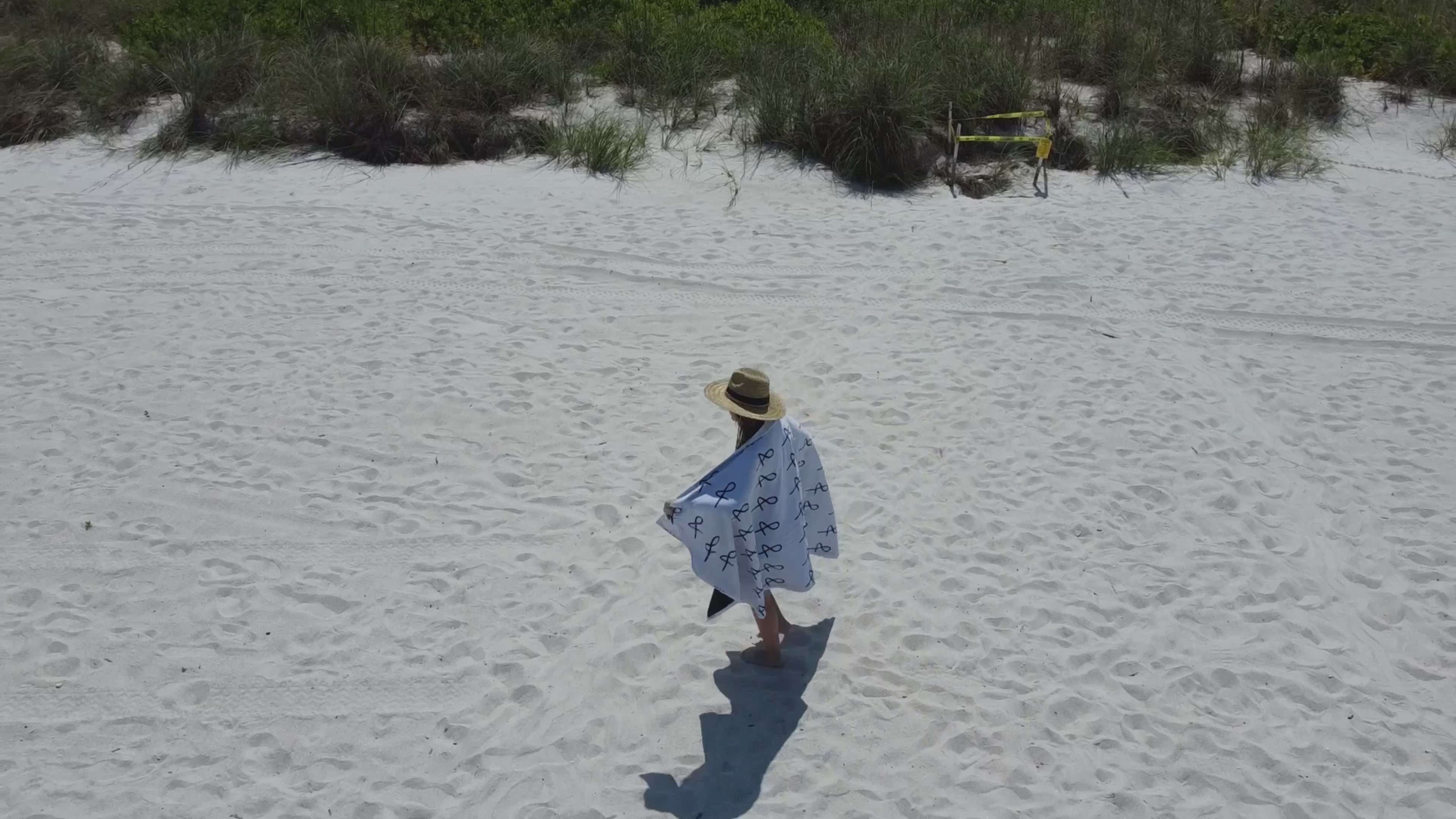 Load video: Person walking along the beach wearing the straw tower hat and A&amp;N beach towel