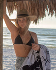 Person under a cabana at the beach wearing the Tower Hat and holding the Anya & Niki beach towel