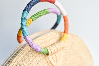Close up of rainbow colored wrapped circle handles on natural raffia straw half-moon clutch with leather sides.