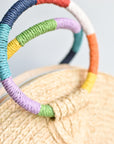 Close up of rainbow colored wrapped circle handles on natural raffia straw half-moon clutch with leather sides.