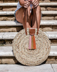 person holding natural seagrass round straw bag with leather handles and suede tassel closure. 