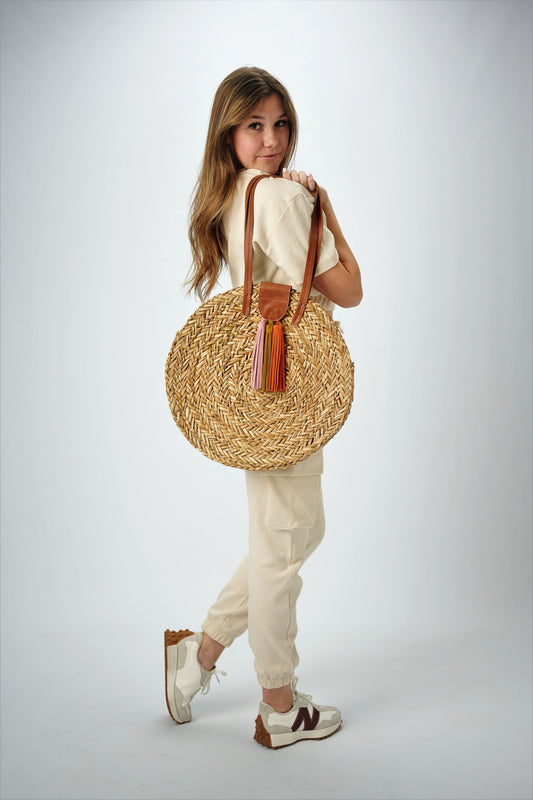 Person holding the Palmdale Straw Bag - a natural seagrass round straw bag with leather handles and suede tassel closure.