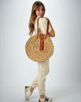 Person holding the Palmdale Straw Bag - a natural seagrass round straw bag with leather handles and suede tassel closure.