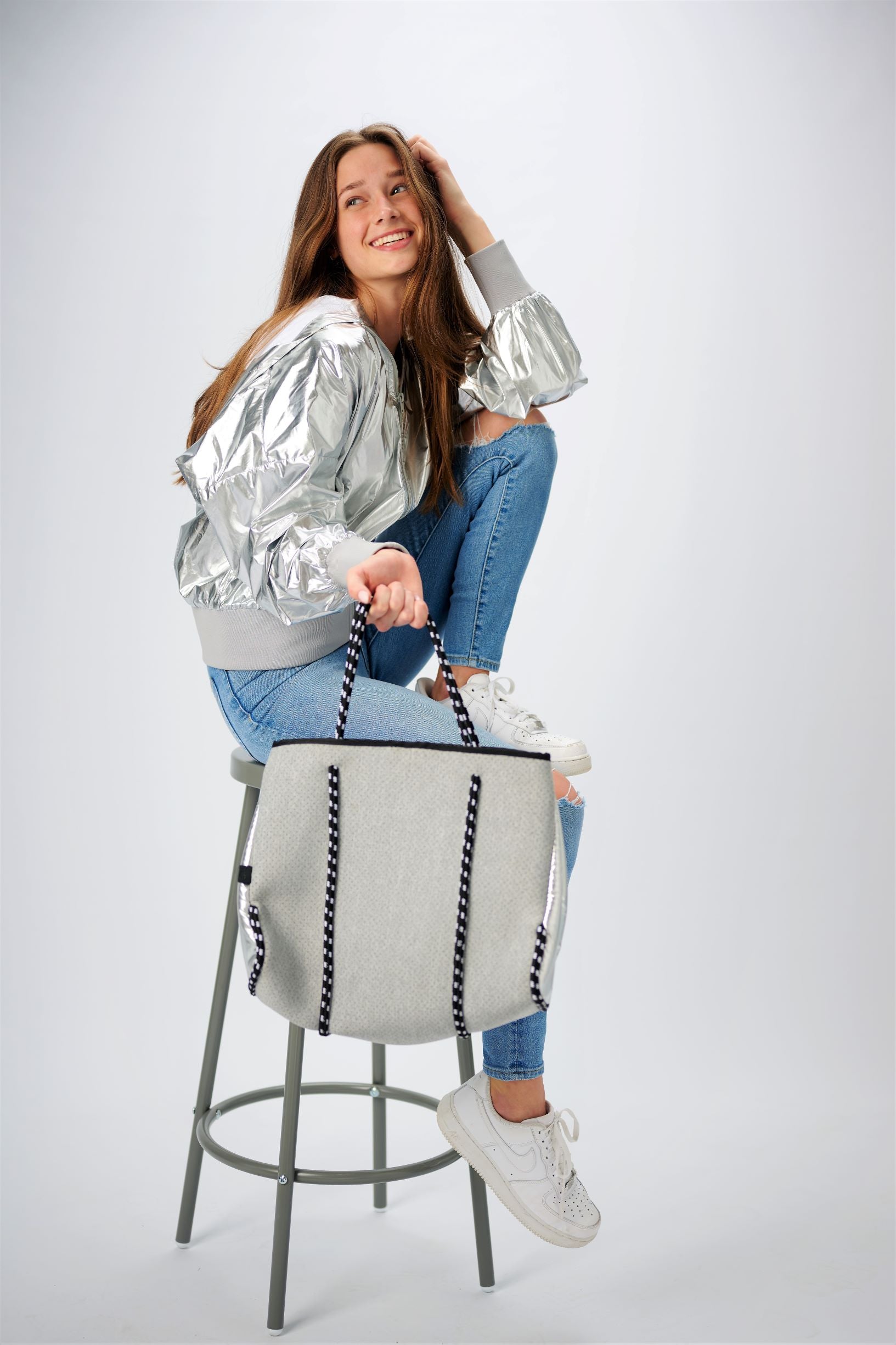 Person sitting on stool holding the Anya &amp; Niki gray neoprene tote bag with shiny silver adjustable sides and cord handles. 