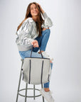 Person sitting on stool holding the Anya & Niki gray neoprene tote bag with shiny silver adjustable sides and cord handles. 