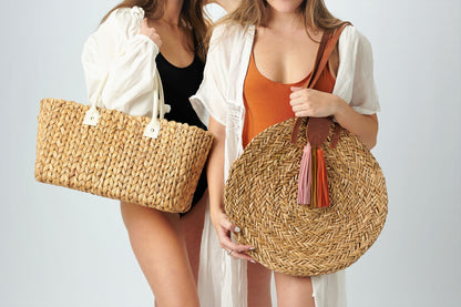 Two people holding Anya & Niki straw bags,  the Augustine hyacinth midsized straw tote with leather handles and the Palmdale circle straw tote with suede tassel detail and leather handles.