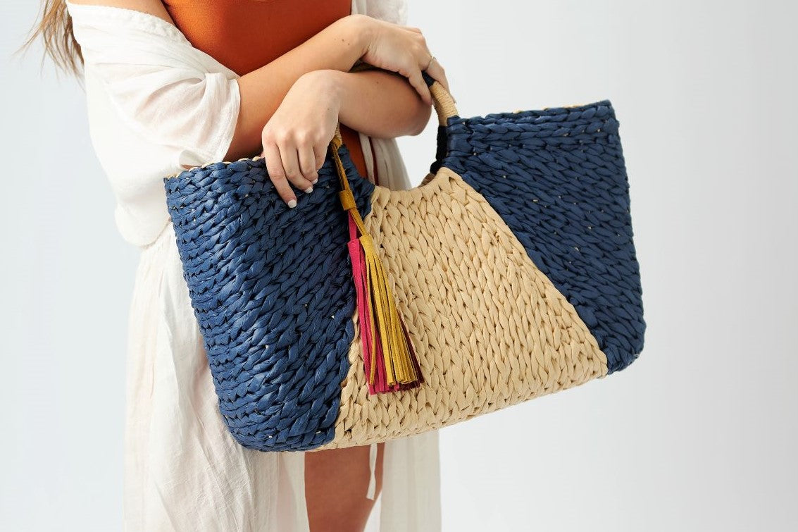 Person holding the Anya & Niki Isla straw bag - a navy and natural colored over-sized straw tote with circle handle and suede tassels.