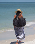 Person at beach holding large black mesh beach bag and wearing the straw Tower Hat with leather chin strap