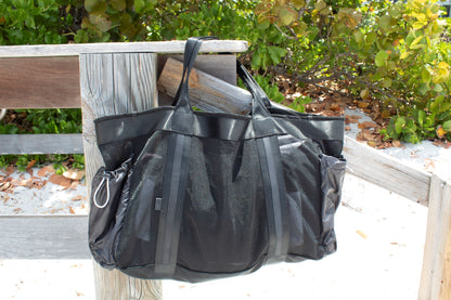 Large black mesh beach bag with nylon side pockets hanging off dock post. 