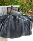 Large black mesh beach bag with nylon side pockets hanging off dock post. 