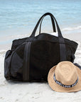 Large black mesh beach bag with nylon side pockets and straw Essential Hat with LOVE band sitting on beach.