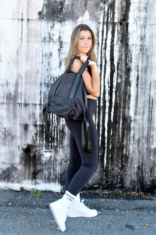 Person holding the Anya & Niki Davie Backpack. A black nylon convertible backpack tote with leather details.