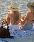 Two people sitting at the beach on the Anya & Niki beach towel, wearing straw hats from Anya & Niki