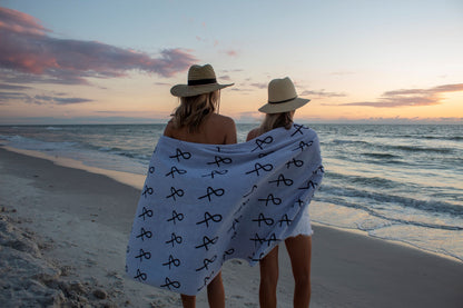 Two people at beach with large Anya & Niki beach towel wrapped around them, wearing straw hats. 