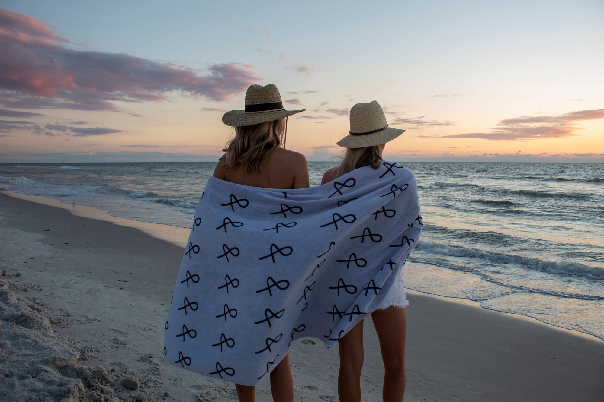 Two people at beach with large Anya &amp; Niki beach towel wrapped around them, wearing straw hats. 