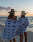 Two people at beach with large Anya & Niki beach towel wrapped around them, wearing straw hats. 