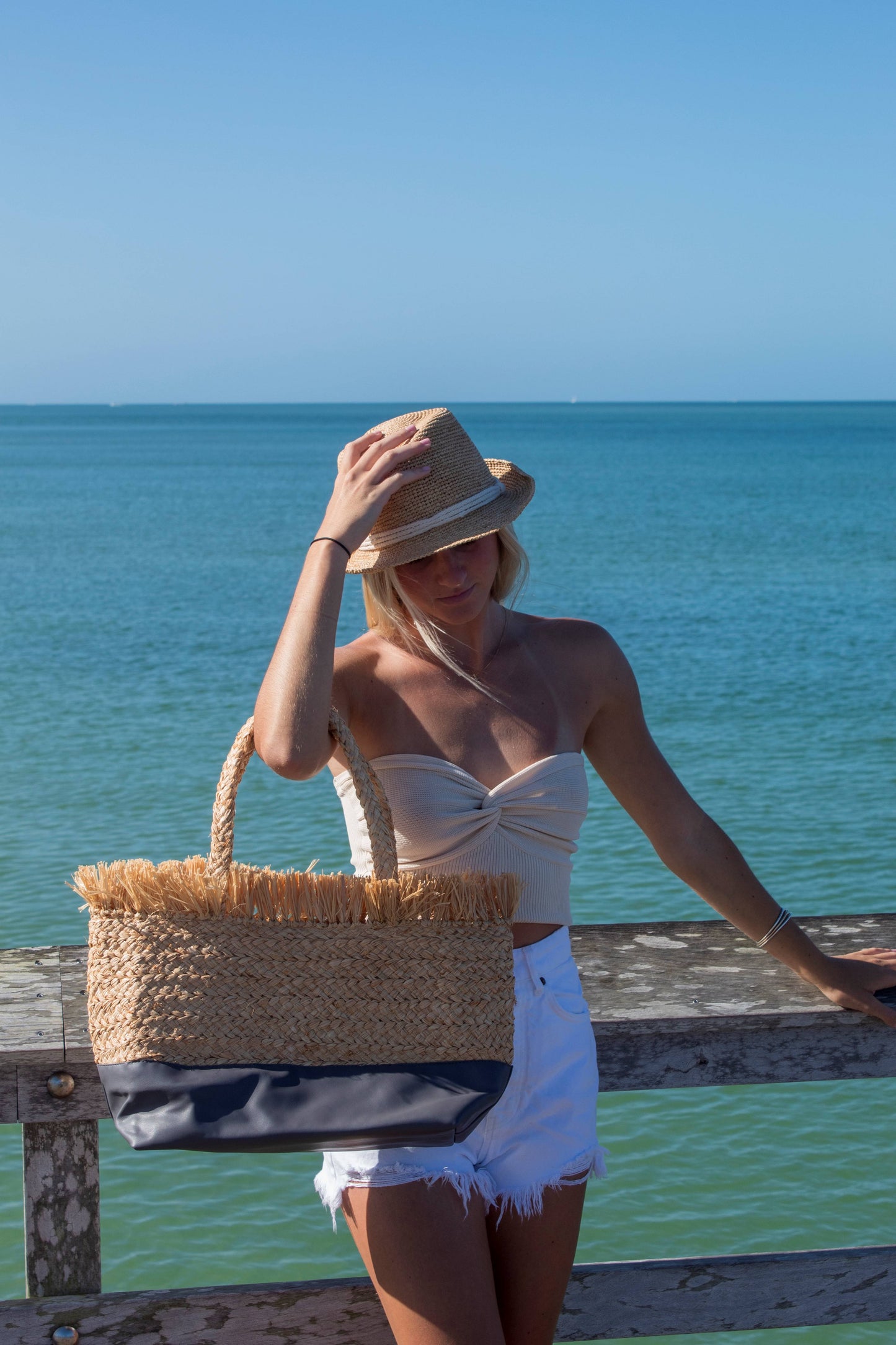 Person at beach holding natural raffia straw tote with linen lining and gray color block base and wearing a raffia straw hat with rope band. 
