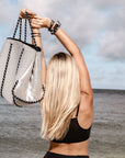 Person at beach holding the Anya & Niki gray neoprene tote bag with shiny silver adjustable sides and cord handles.